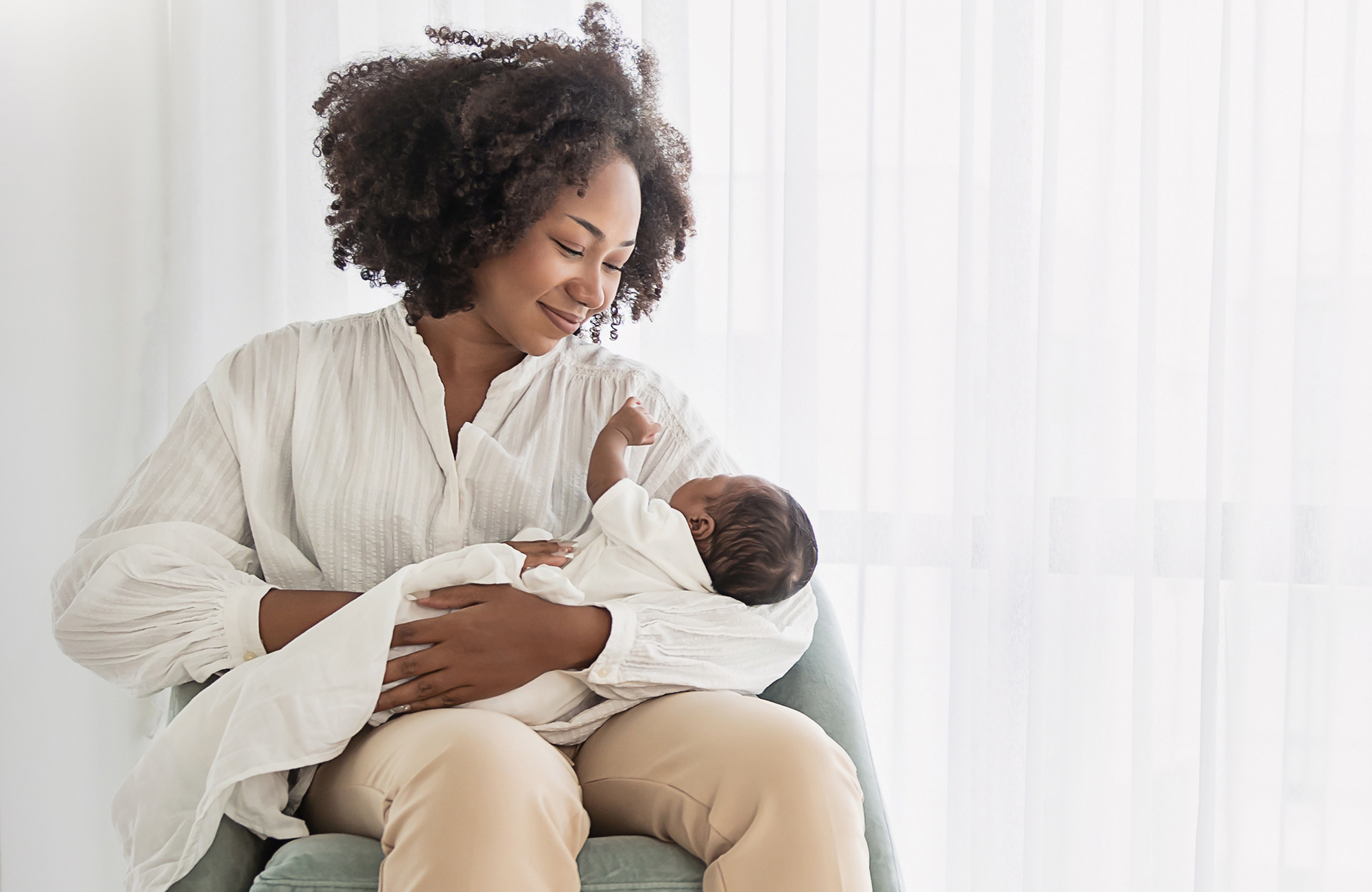 A mother holds her baby in her arms while she's sat down, she's smiling at the baby
