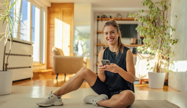 A woman is sat smiling at the camera on her yoga mat with a phone in her hands