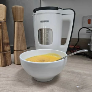 A bowl of fresh soup sits next to the Morphy Richards Total Control soup maker atop a wooden surface