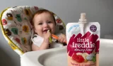 A baby in a highchair with a spoon in their mouth, in the forefront is a Little Freddie pouch