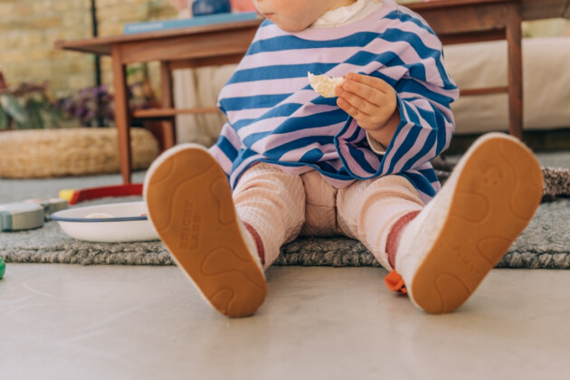 A toddler is sat on the floor having a snack, feet facing the camera