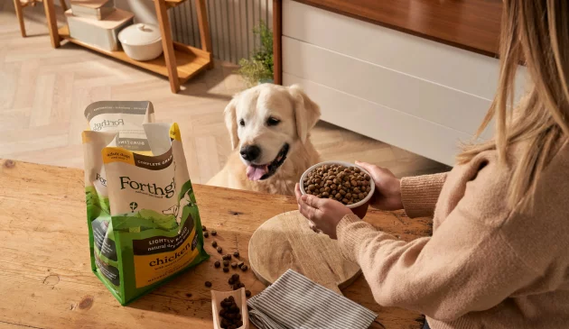 A dog sits next to its owner at the table as she holds his dog bowl filled with Forthglade food