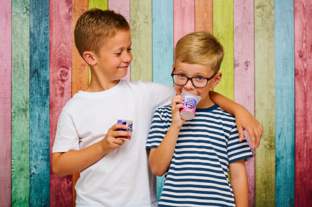 Two boys eating a Petits Filous yoghurts, one with his arm around the other, against a rainbow wall