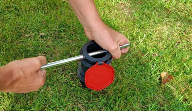 A pair of male hands using a metal rod to screw the plastic ground screw into grass