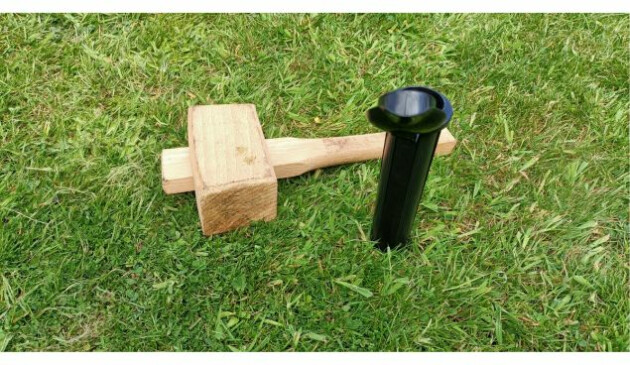 A black plastic ground socket for a rotary airer partially knocked into the ground. A wooden mallet lays on the grass next to it.