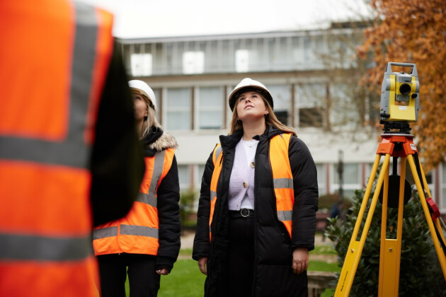 A further education student stands on a surveying site with a high vis and helmet on next to a camera