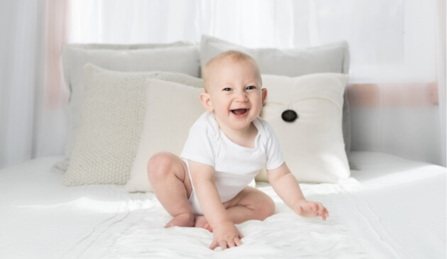 Baby boy dressed in white vest smiling and sitting on bed