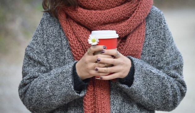 A close up of a women's hands and mid section. She has purple nails and various rings on her fingers and is holding a red paper coffee cup with a white plastic lid and a white and yellow daisy. She is wearing a thick grey woollen jumper and a thick rust coloured woollen scarf