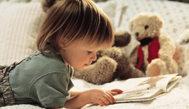 A child lying on a bed, reading a book. There's a teddy bear in the background of the image.