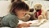 A child lying on a bed, reading a book. There's a teddy bear in the background of the image.