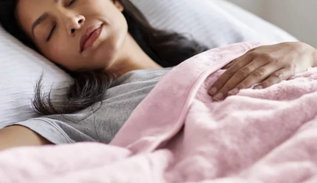 A woman sleeps in bed with a Cosi Home heated throw over her