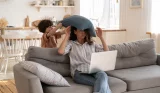 A mum is hit on the head with a pillow by her child while she tries to work on a laptop