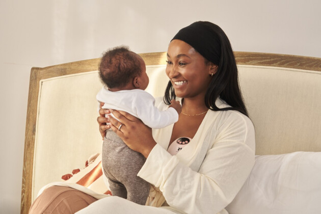 A mother lays in bed and is holding her baby while pumping