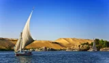 A sail boat floating on the river with sandy hills and trees in the background
