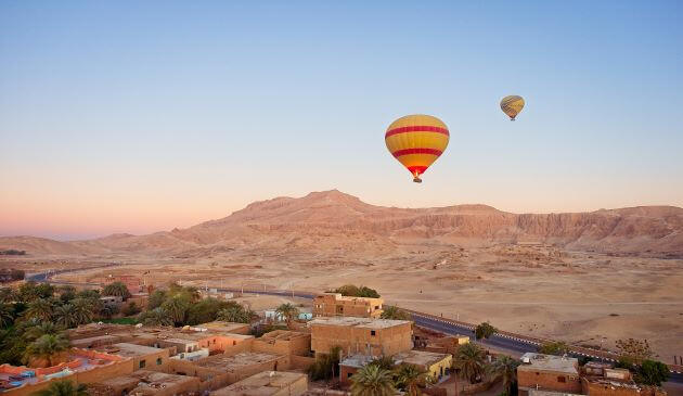 Image of desert, mountains and cement huts wuth some palm trees and two yellow and red hot air balloons in the sunset sky..