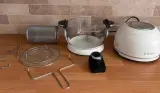 White Fritaire glass bowl air fryer with its lid, tumbler basket, air stand, rotisserie parts, motor and grabber laid out on a kitchen worktop