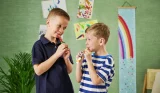 Two young boys look at each other as they eat Frube yoghurts against a green wall