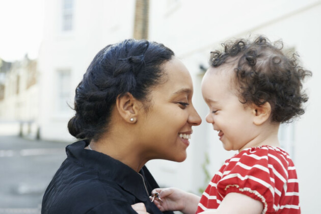 A mother smiles while holding her baby who is facing her, they're stood outside