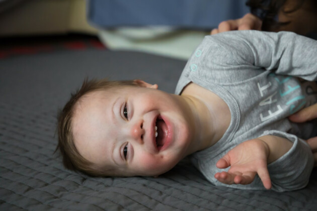 A baby with Down Syndrome smiles at the camera while being tickled on the floor