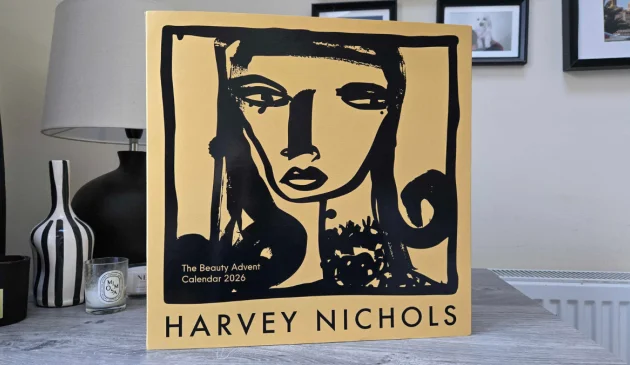 Front of the Harvey Nichols Beauty Advent Calendar 2026 sleeve, in yellow with a black brushstroke illustration of a woman’s face, standing on a grey wooden table beside a lamp, a striped vase and candles
