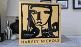 Front of the Harvey Nichols Beauty Advent Calendar 2026 sleeve, in yellow with a black brushstroke illustration of a woman’s face, standing on a grey wooden table beside a lamp, a striped vase and candles