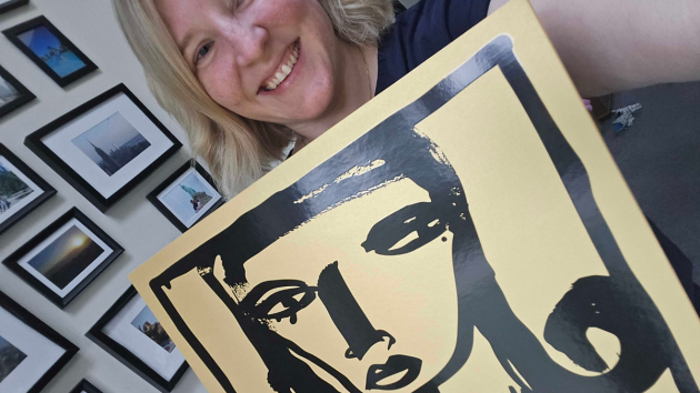 A smiling woman with blonde hair holding the glossy black face illustration on the yellow calendar sleeve up to the camera, with a wall of framed photos behind her