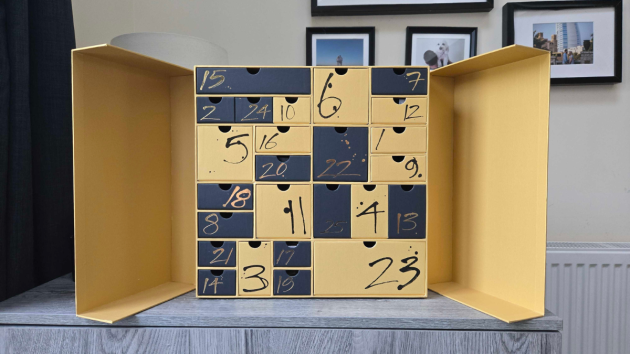 The calendar with both doors open, showing 25 drawers of different sizes in yellow and navy, each with a hand-painted number in black or gold