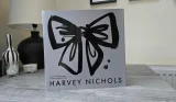 Front of the Harvey Nichols 12 Days of Fragrance Advent Calendar 2026 sleeve, in silver-grey with a large black brushstroke bow, standing on a grey wooden table beside a lamp, a striped vase and candles
