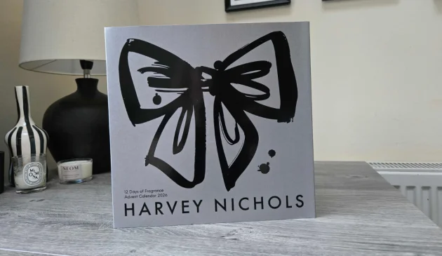 Front of the Harvey Nichols 12 Days of Fragrance Advent Calendar 2026 sleeve, in silver-grey with a large black brushstroke bow, standing on a grey wooden table beside a lamp, a striped vase and candles