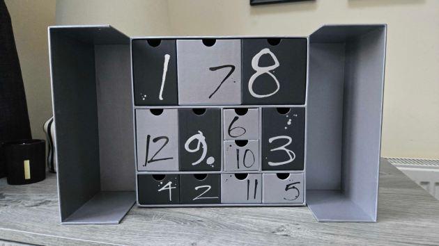 The calendar with both doors open, showing 12 drawers in dark and light grey in different sizes, each with a hand-painted number in white or black