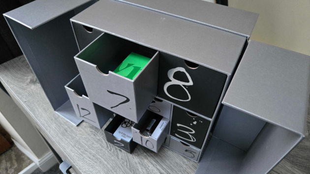 Angled view of the open calendar with several drawers pulled out, showing a green box inside drawer 7 and fragrance boxes in drawers 2 and 10