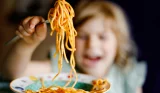 A little girl holds up spaghetti to the camera, she's out of focus but smiling in the background