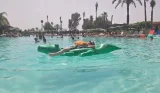 Child wearing an orange life jacket relaxes on a green crocodile inflatable in the hotel’s busy main pool.