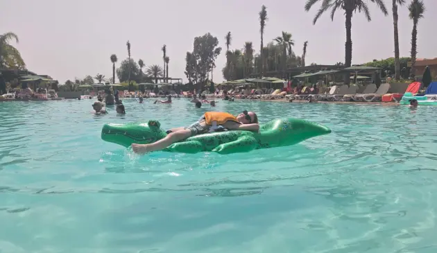 Child wearing an orange life jacket relaxes on a green crocodile inflatable in the hotel’s busy main pool.