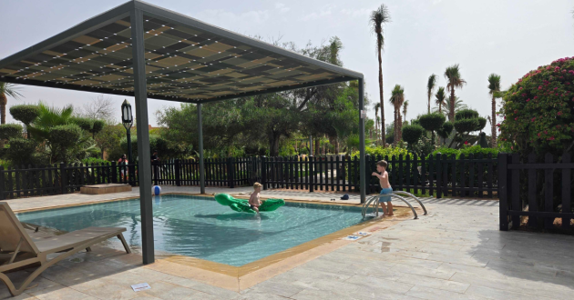 Two children play with a green crocodile inflatable in a small fenced swimming pool beside shaded loungers