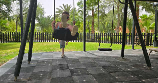 One child sits in a bucket swing while another pushes from behind in the hotel’s shaded playground