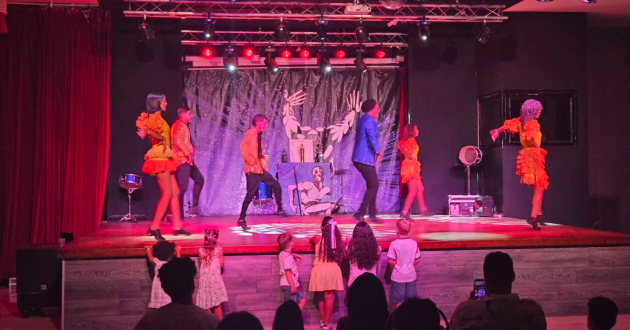 Hotel entertainers dance on a brightly lit indoor stage while children watch from the front of the audience