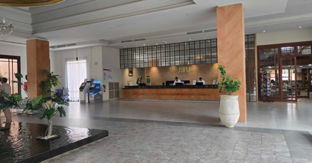 Hotel reception desk beneath a gold RIU sign, with an ATM, plants and the gift shop nearby