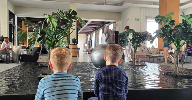 Two children sit beside a shallow indoor water feature surrounded by large tropical plants in the hotel lobby