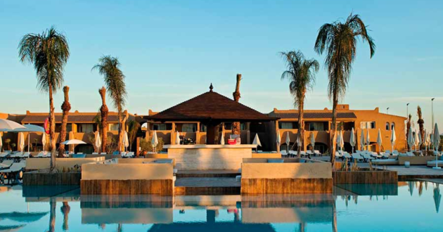 Main swimming pool and central pool bar surrounded by sunloungers, parasols, palm trees and terracotta buildings