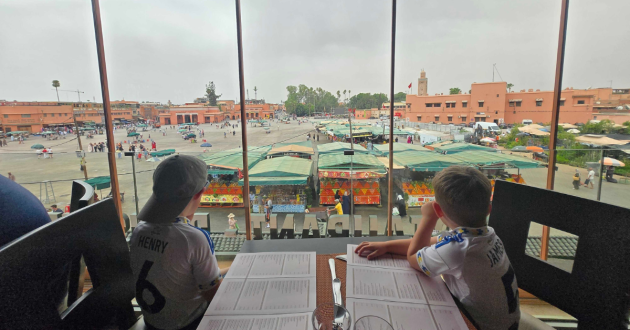 Two children sit at a restaurant table overlooking the market stalls and terracotta buildings of Jemaa el-Fnaa.