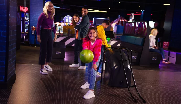 A young girl is smiling while holding a bowling ball with her family stood cheering behind her