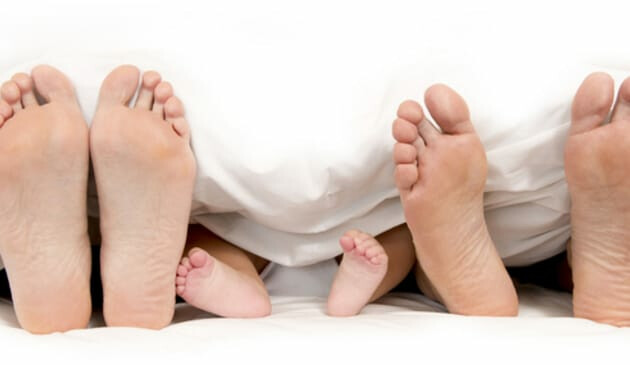 Two pairs of adult feet and one pair of baby feet sticking out of the end of a bed