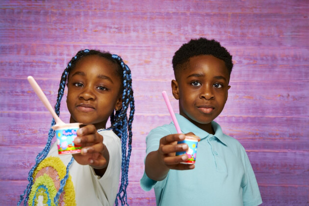 Two children hold up their Petits Filous yoghurts to the screen as they stand against a purple wall