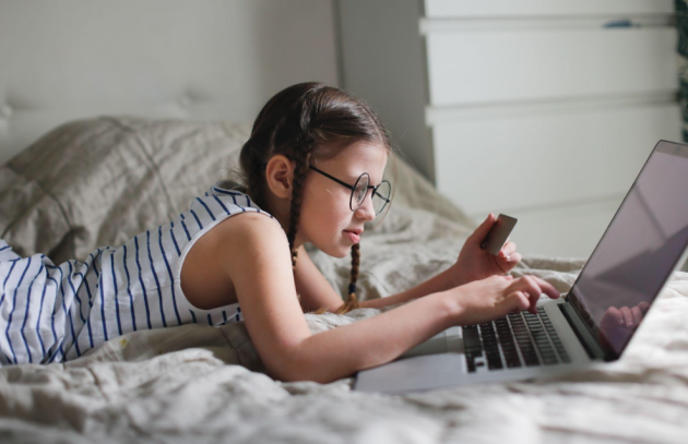 A young girl using her GoHenry debit card to pay for something on her laptop while laid on her bed