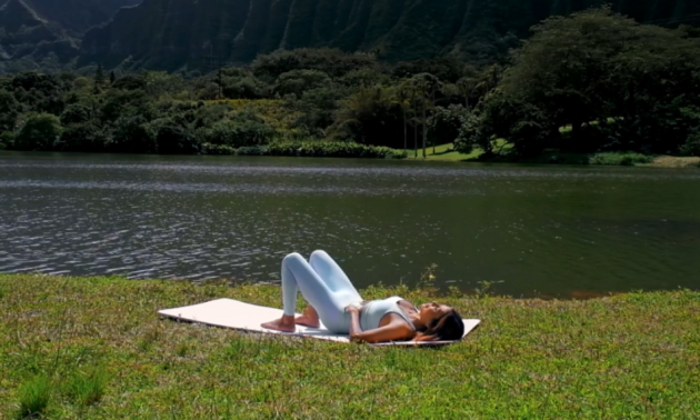 A woman does pelvic tilts on a yoga mat on the grass next to water