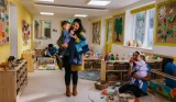 Nursery worker holding young child while parents and children play together in cheerful daycare classroom with natural toys