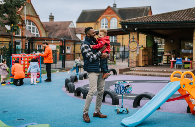 Father holding toddler at outdoor nursery playground with colorful equipment and tire obstacles in English village setting