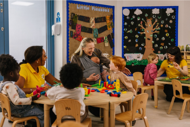A mum holds her baby in a sling wrap while playing with her older child at nursery