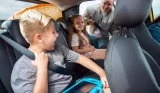 A brother and sister smile at each other in the back of a car while their mum checks on them